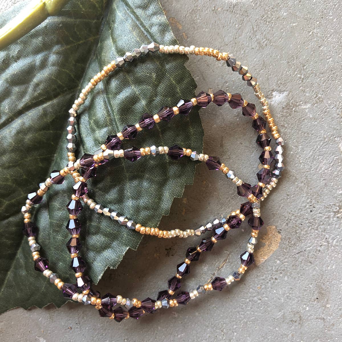 Close-up of purple and gold Nourish Cause Connection handmade beaded bracelet displayed on a green leaf.