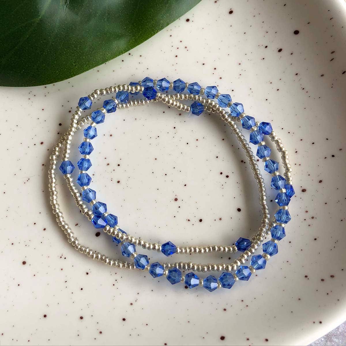 Two blue handmade beaded bracelets by women artisans on a speckled surface with a green leaf