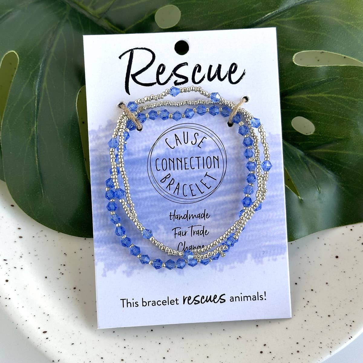 Blue beaded bracelet on a 'Rescue Cause Connection Bracelet' card with green leaves in the background