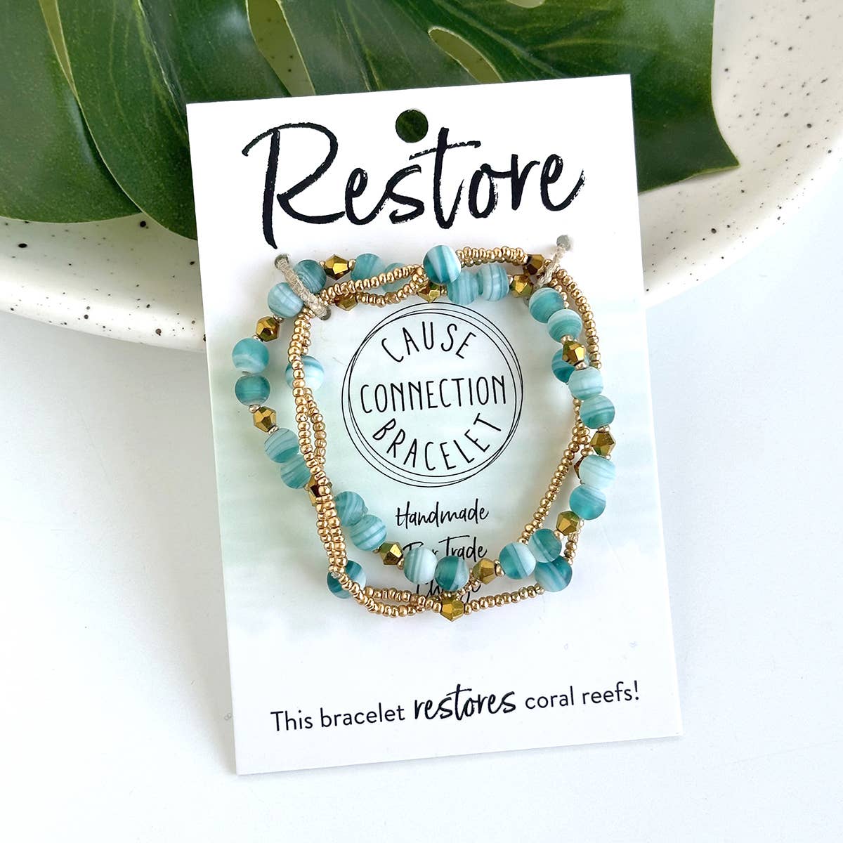Close-up of turquoise and gold handmade beaded Restore bracelet on a white card, a fair trade bracelet that helps restore coral reefs.